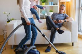 Frau mit Kind auf dem Arm saugt Staub während Mann auf dem Sofa sitzt und Handy nutzt